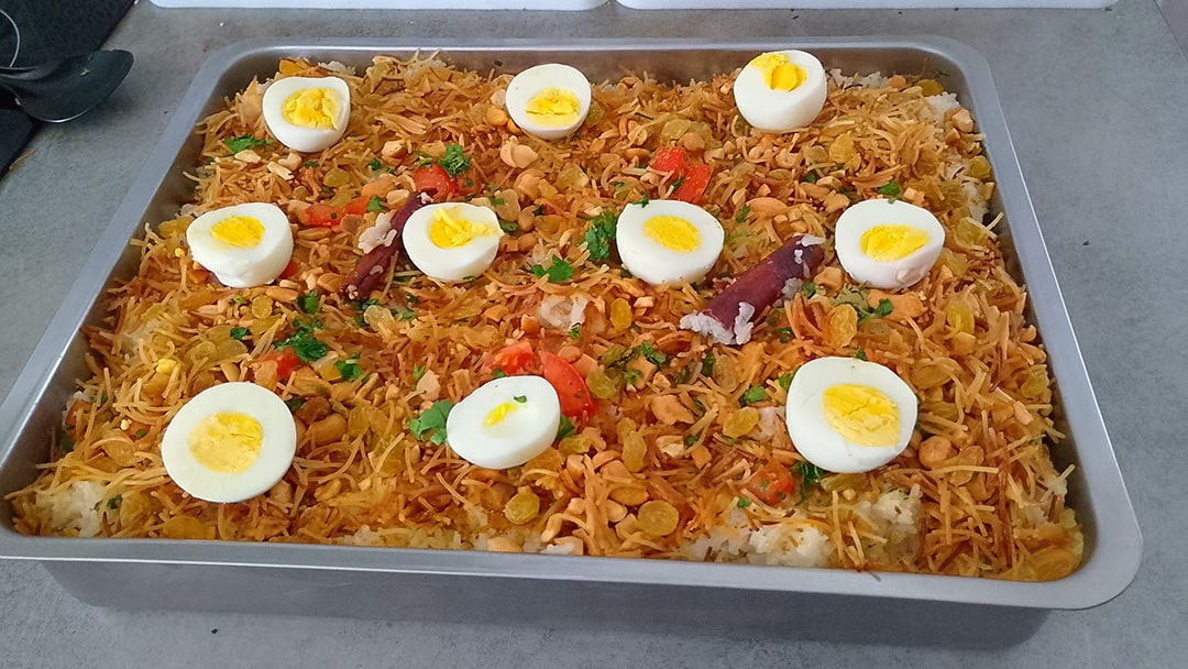 Plat de biryani au riz basmati parfumé aux épices, garni de morceaux de poulet, œufs durs, oignons caramélisés, vermicelles dorés, servi avec du yaourt grec et une sauce antchari maison